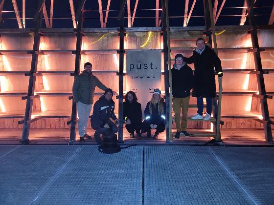 The Haugland group (Marius, Danijel, Mateusz, Floriane, Stian, Anna) outside the uplit facade of the Pust sauna.