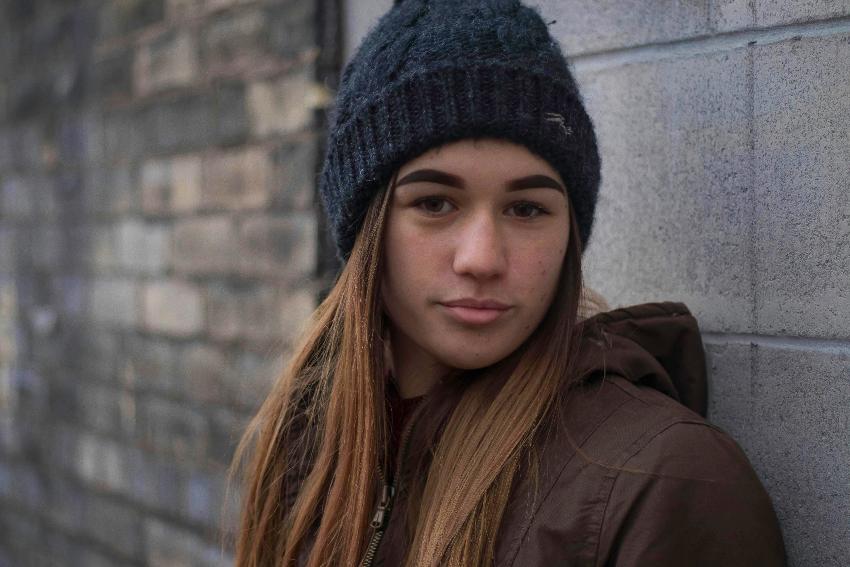 Ung jente med langt h&aring;r lener seg mot en vegg og ser mot kamera