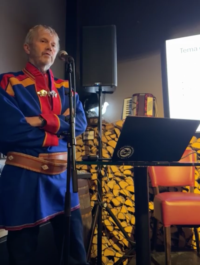 mann, har armene i kors, har p&aring; samisk drakt, snakker i mikrofon, st&aring;r p&aring; en scene med teknisk utstyr rundt.