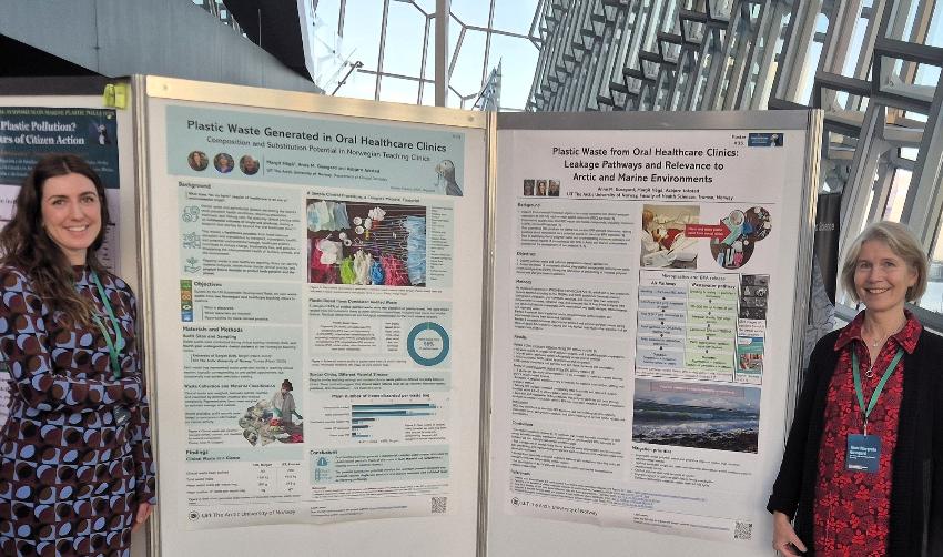 To kvinner st&aring;r foran poster og smiler til kamera